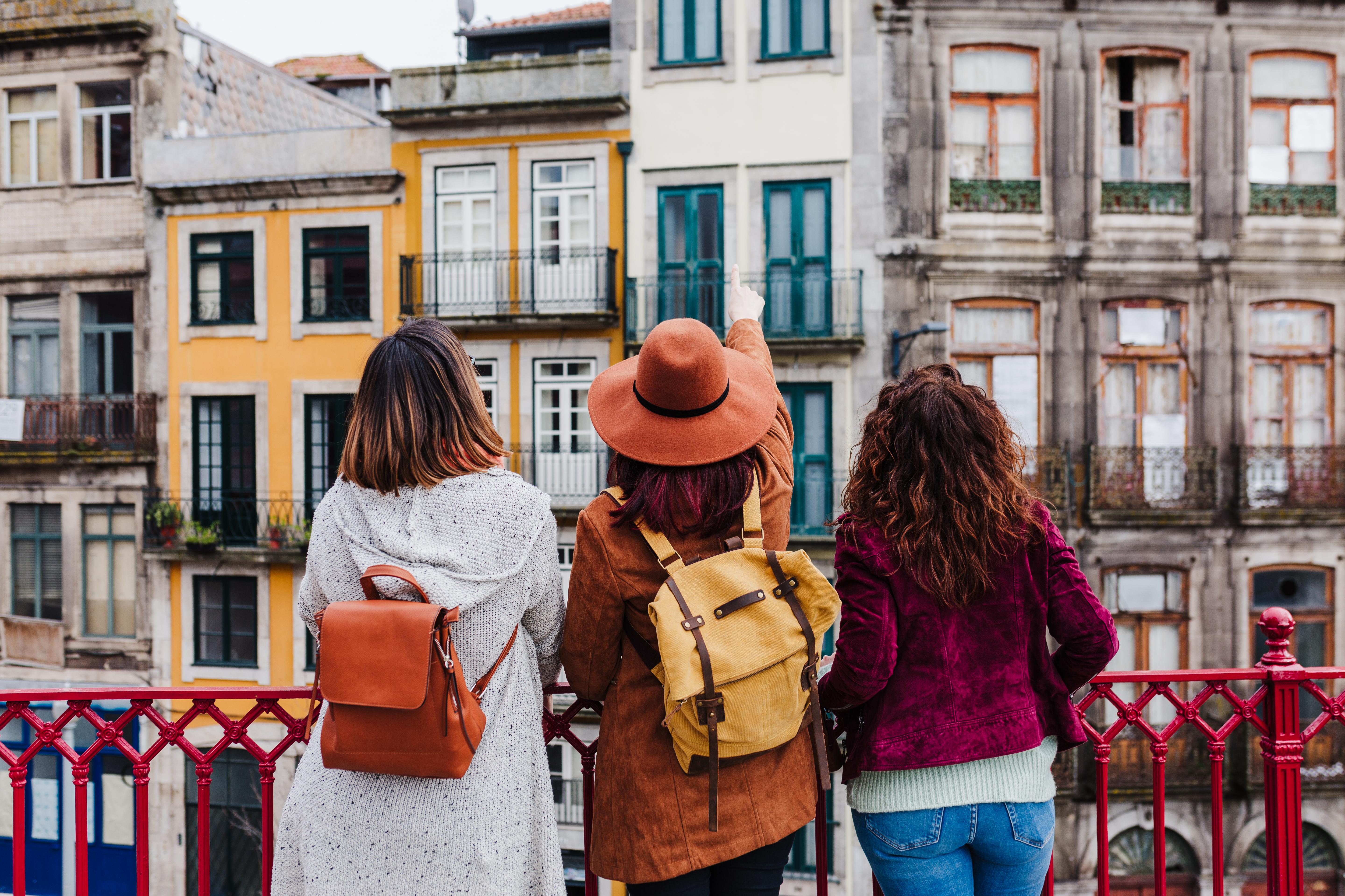Três viajantes observam edifícios históricos numa rua do Porto, vistas de costas, com mochilas às costas e varandas tradicionais ao fundo.