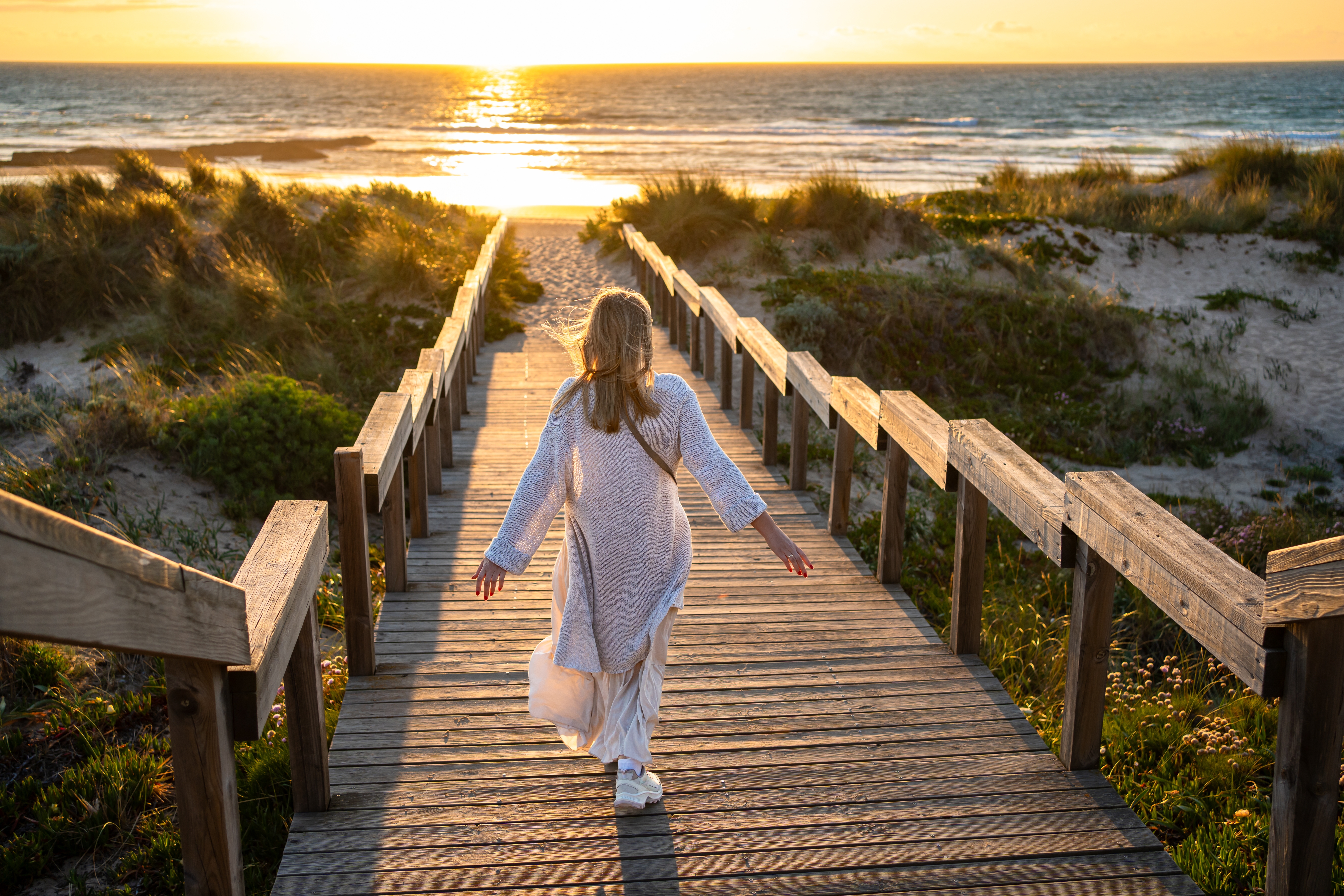 Pessoa a caminhar num passadiço de madeira em direção à praia ao pôr do sol em Portugal