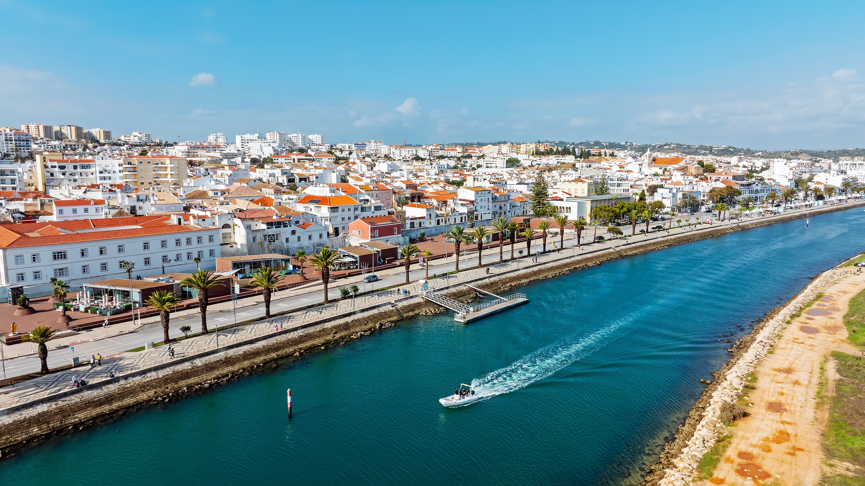 Vista aérea de Lagos com a Ribeira de Bensafrim em primeiro plano, passeio ribeirinho com palmeiras e embarcação a navegar.